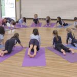 grupa dzieci w sali szkolnej wykonuje pozę drzewa z uniesionymi rękami pod okiem nauczycielki