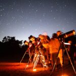 grupa dzieci i dorosłych obserwuje gwiaździste niebo przez teleskopy na ciemnej łące