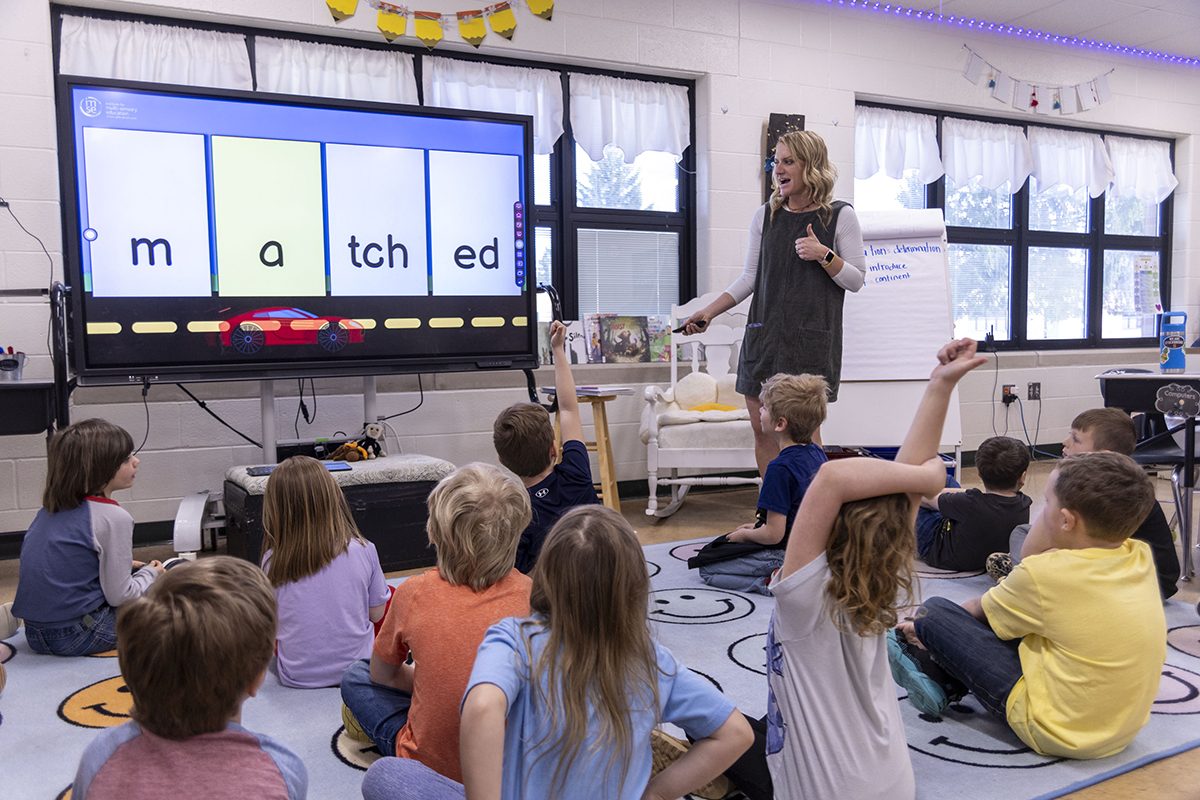 Nauczyciel wskazuje na kolorowy slajd na ekranie przed grupą skupionych uczniów w jasnej klasie